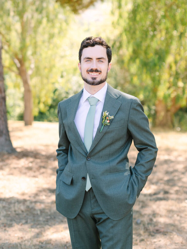 A smiling groom in a green suit with a matching tie stands outdoors in a sunny park, hands in pockets, exuding a cheerful, relaxed demeanor.