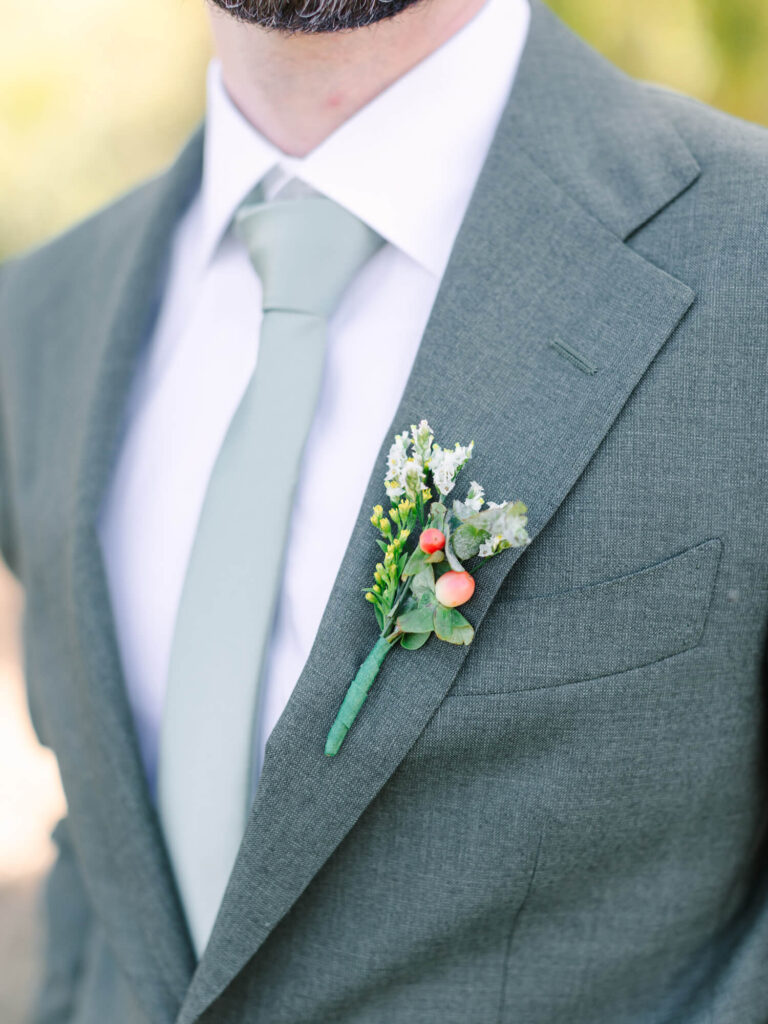 A man wears a sage green suit with a matching tie and a boutonniere of white flowers and red berries.