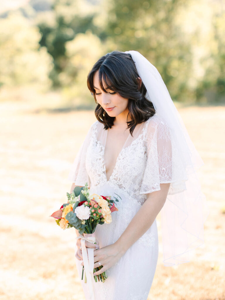 A bride in a white lace wedding dress holds a colorful bouquet, gazing down softly.