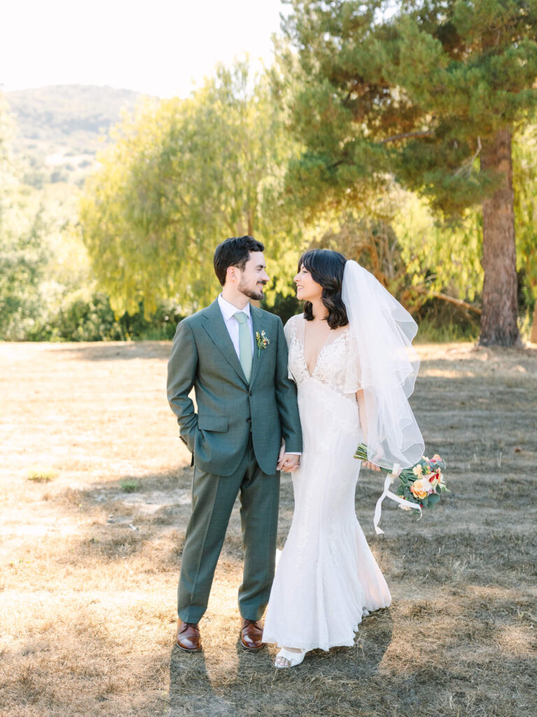 Bride and groom stand smiling in a sunlit park, the bride in a white dress holding flowers, the groom in a green suit.