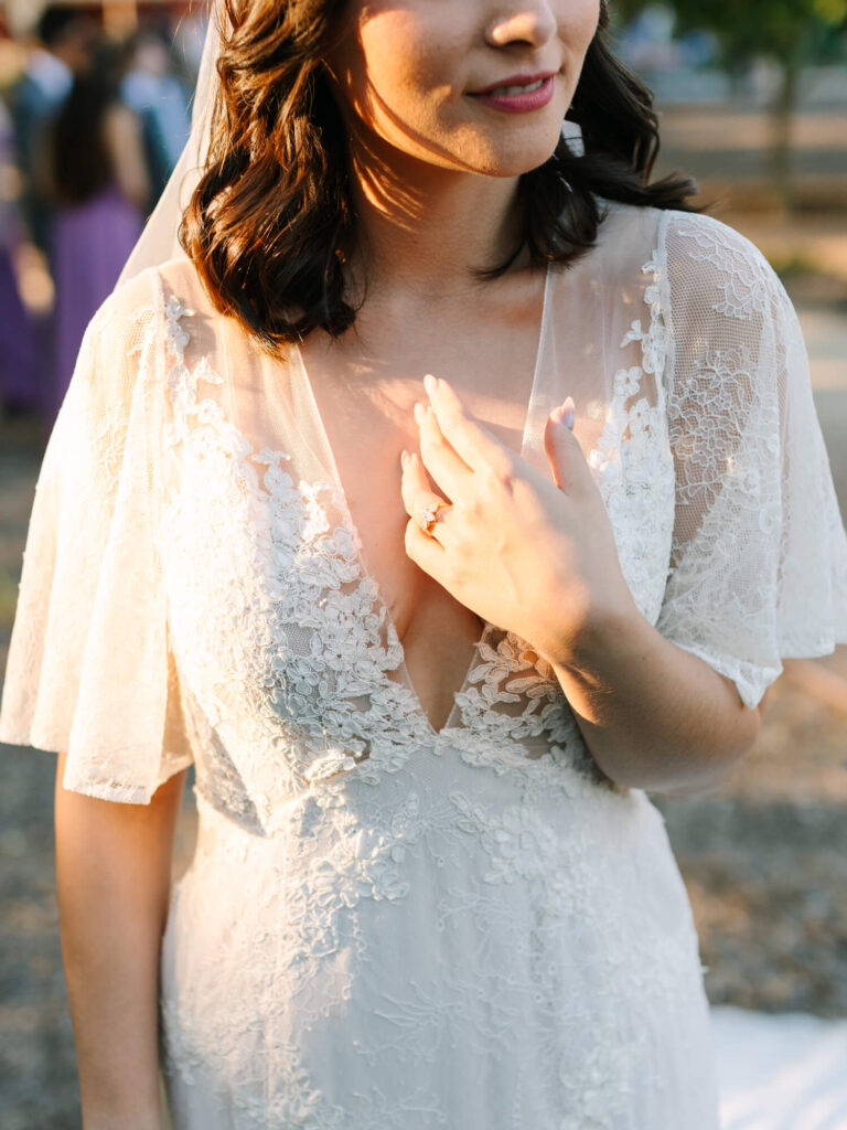 Bride in sunlight wearing a lace wedding gown, holding her chest with left hand.