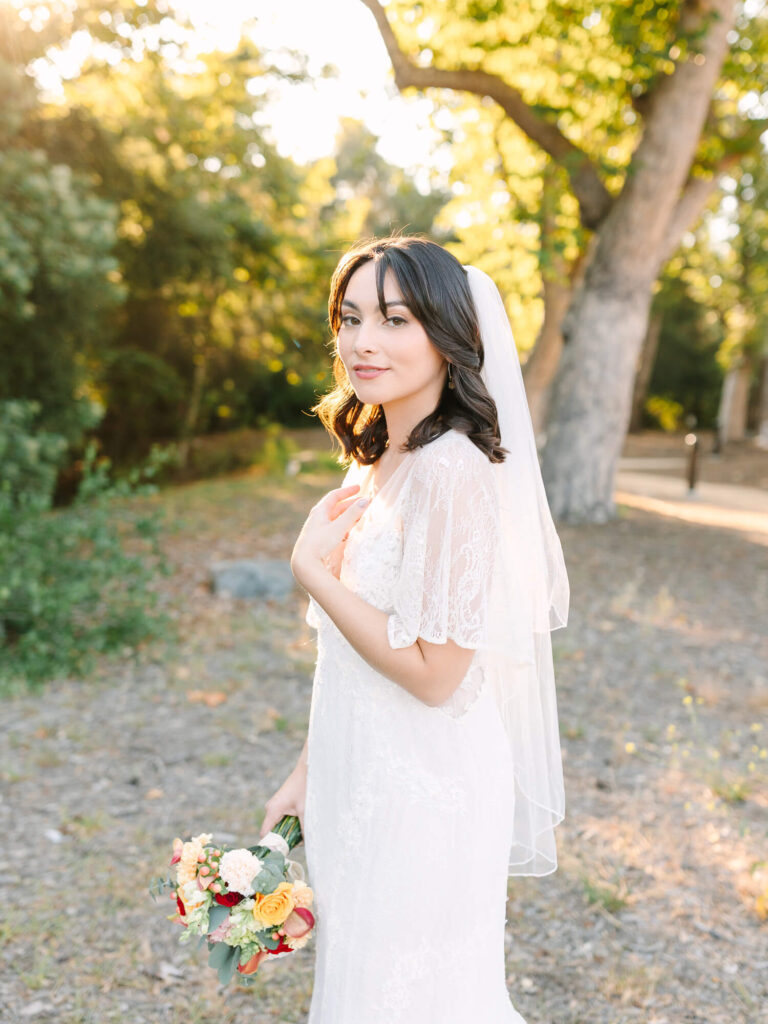 Bride in lace gown and veil, holding a bouquet with red and yellow flowers.