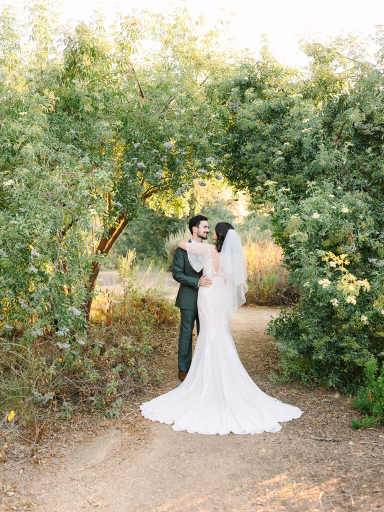 A bride and groom embrace under a lush green archway. The bride wears a flowing white gown and veil.