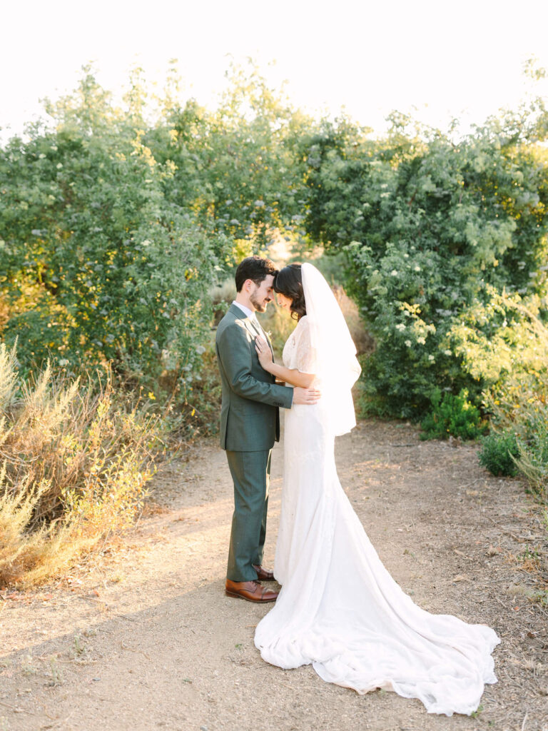 A bride in a white dress and veil, and a groom in a suit, stand on a sunlit path surrounded by lush greenery, sharing an intimate moment.