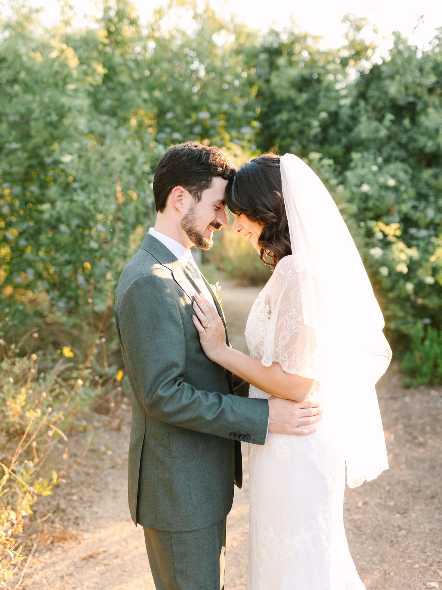 A bride in a lace dress and veil embraces a groom in a green suit on a sunlit forest path. They touch foreheads, smiling tenderly.