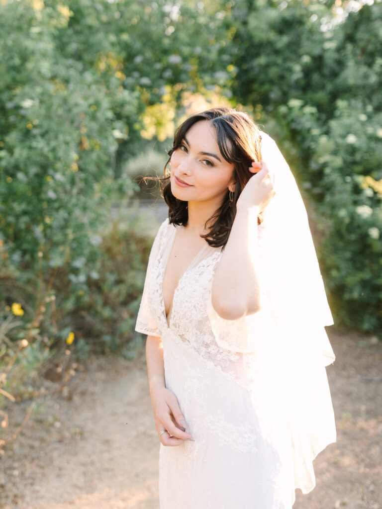 A bride in a lace wedding dress and veil stands on a sunlit path, surrounded by lush greenery. She gently touches her veil, smiling softly.