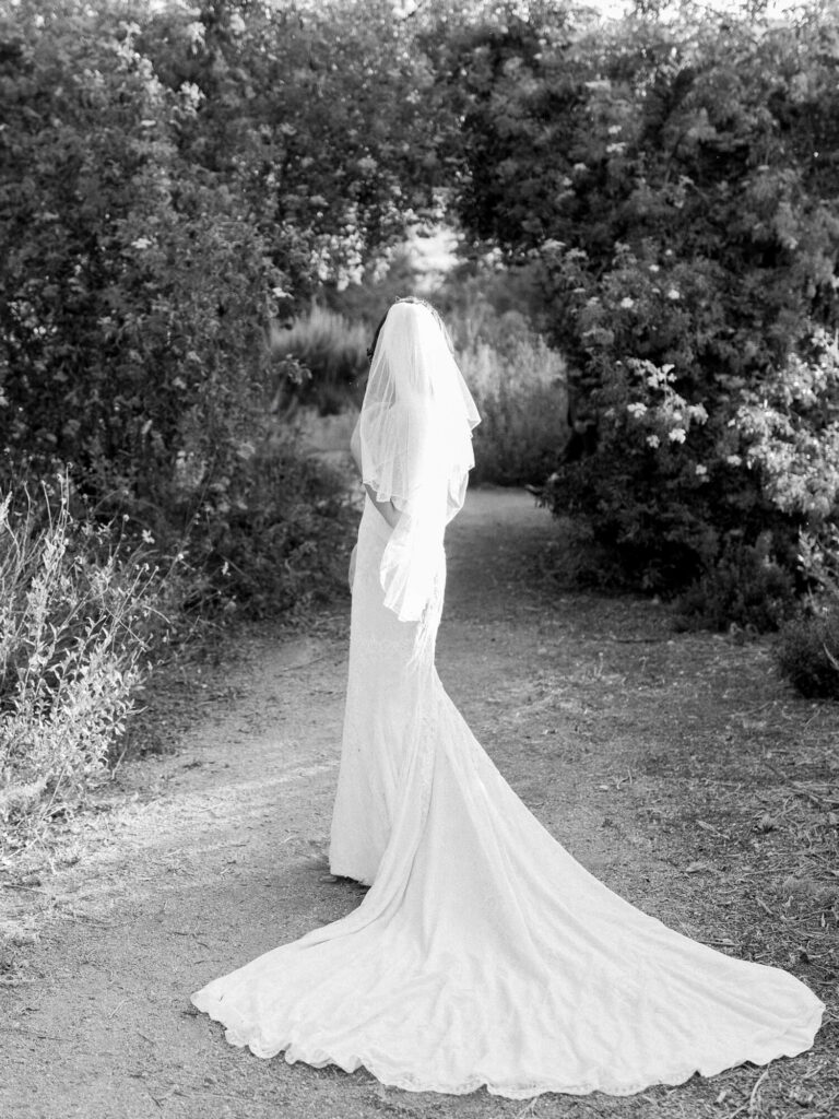 A bride in a long, flowing gown and veil stands on a path surrounded by lush foliage.