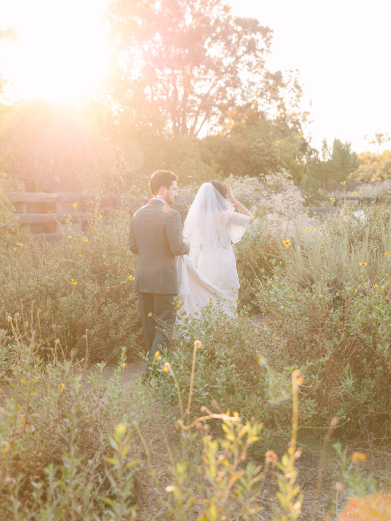 Bride and groom walk through a sunlit field, surrounded by wildflowers with a golden sunset in the background.