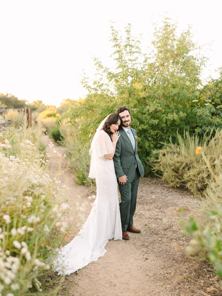 Bride in a flowing white dress leans affectionately on groom in a green suit.