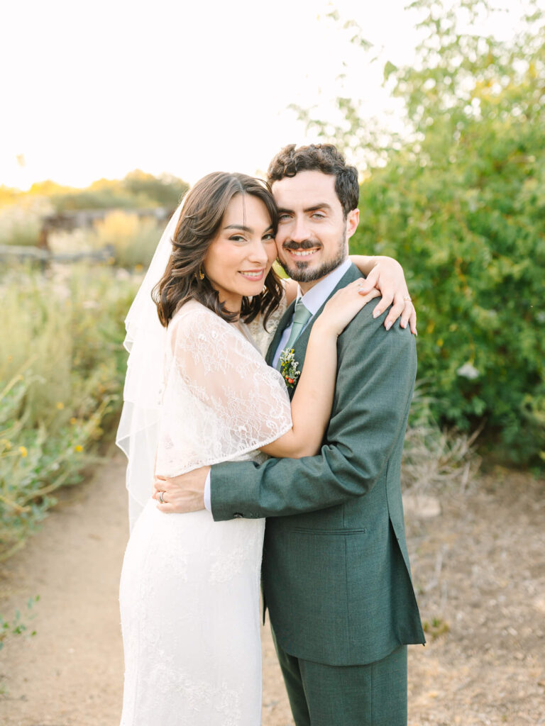 A smiling couple in wedding attire embraces on a rustic path surrounded by lush greenery.
