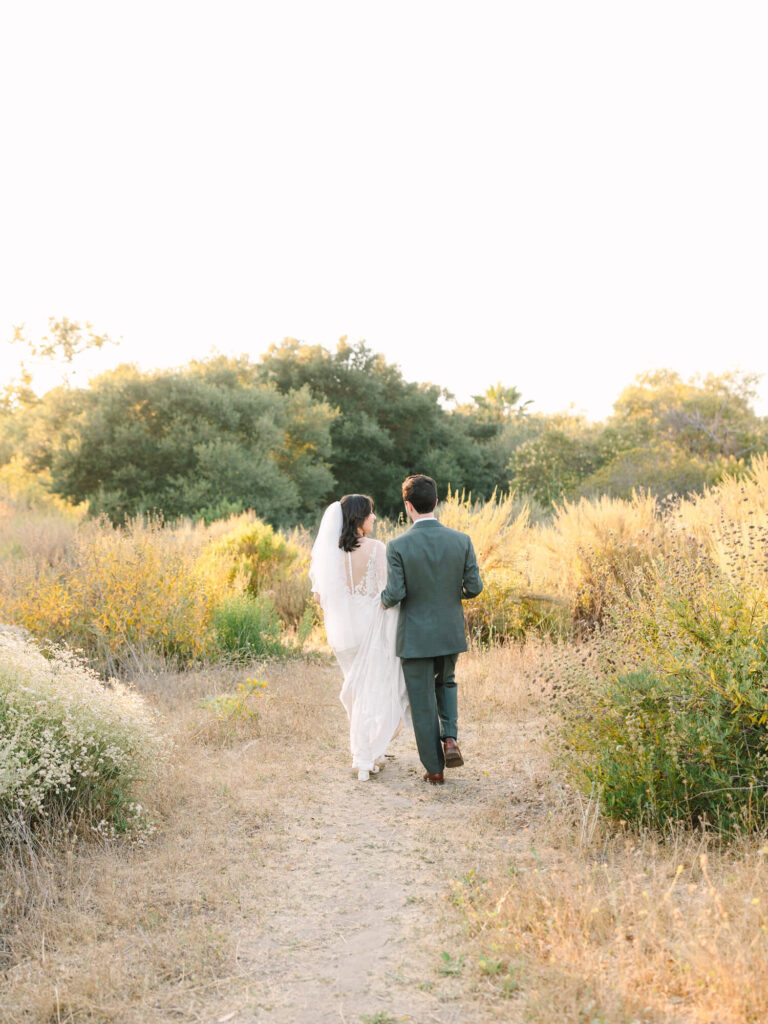 Bride and groom walking on a sunlit path, surrounded by tall grass and trees.