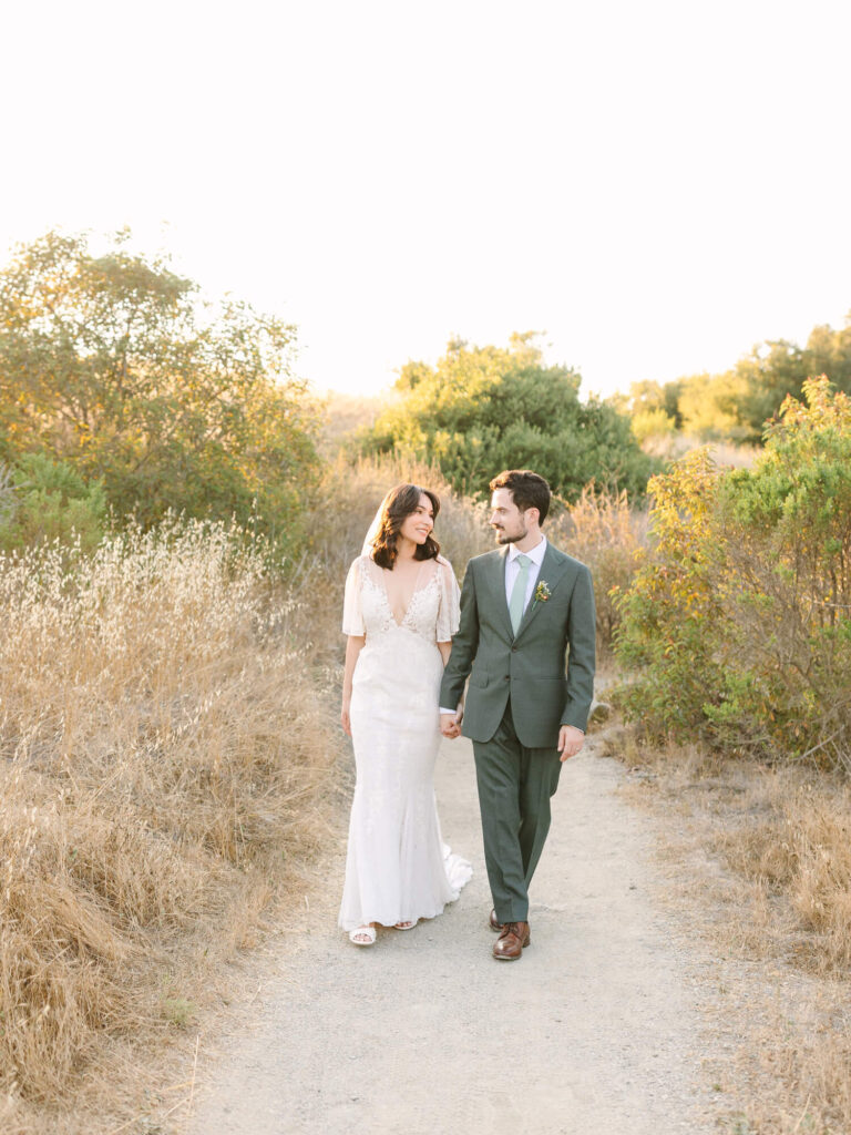 Bride in a white lace gown and groom in a green suit walking hand in hand on a sunlit dirt path, surrounded by golden fields and shrubs.