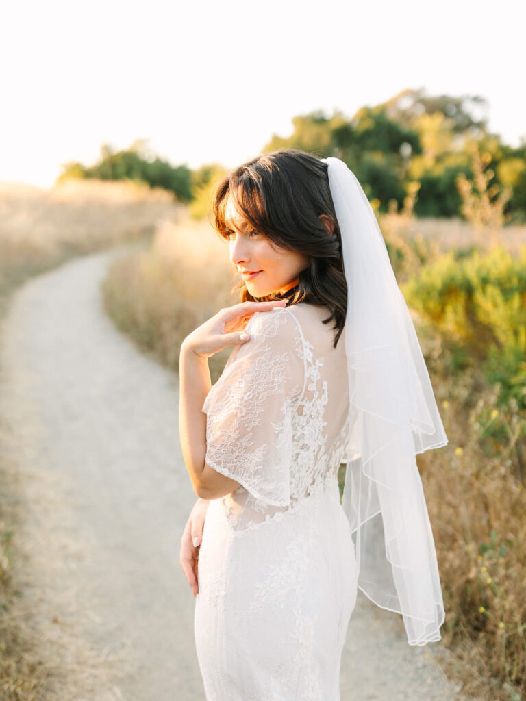 A bride in a lace wedding dress and veil stands on a sunlit path, surrounded by nature. She gazes softly over her shoulder.
