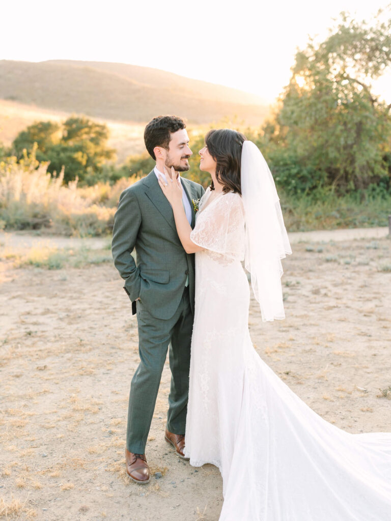 Bride in lace dress and veil embraces groom in a green suit in a sunlit field gazing at each other lovingly.