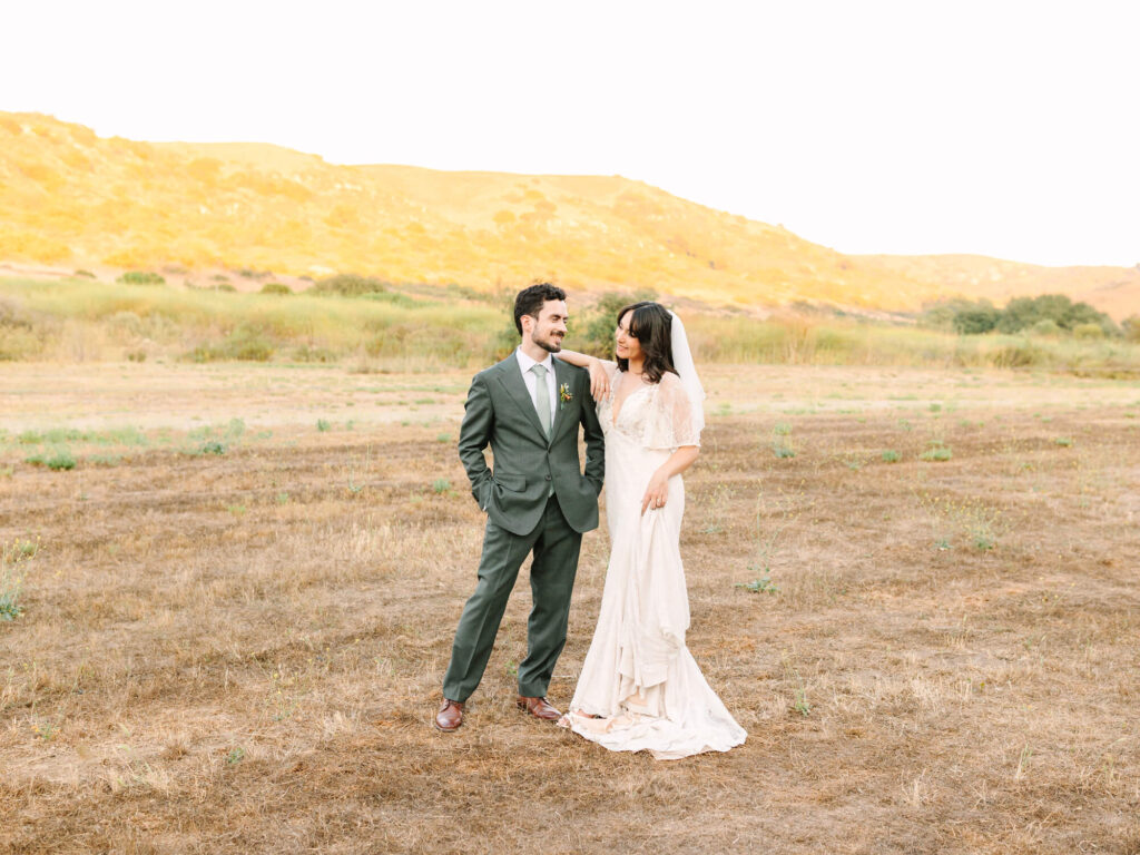 A bride in a flowing white dress stands with her arm around a groom in a green suit against a sunlit, grassy landscape.