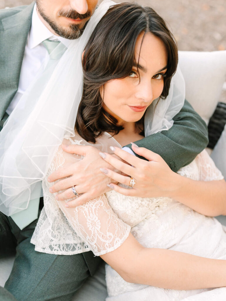 Bride in lace dress and veil, embraced by groom in green suit, both displaying rings.