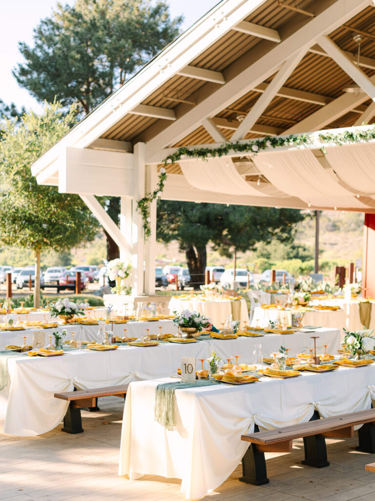 Elegant outdoor wedding reception under a white pavilion, featuring long tables with white cloths, yellow tableware, and greenery.