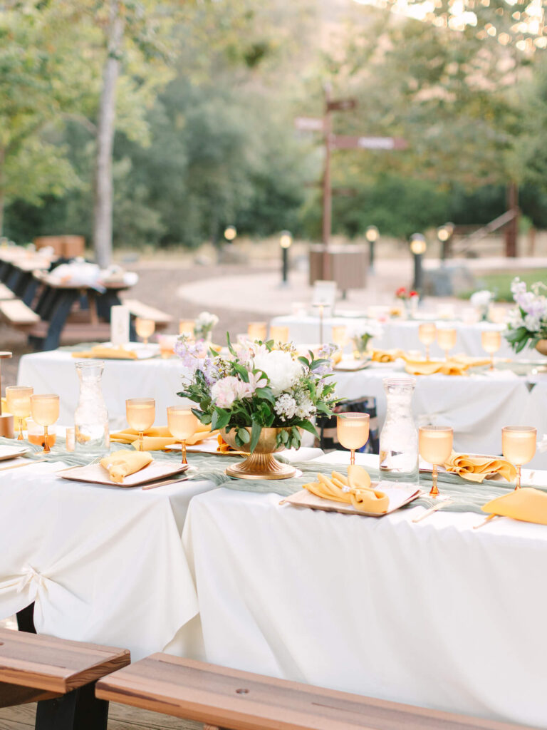 Outdoor dining setup with elegant white tablecloths, floral centerpieces, amber goblets, and yellow napkins.