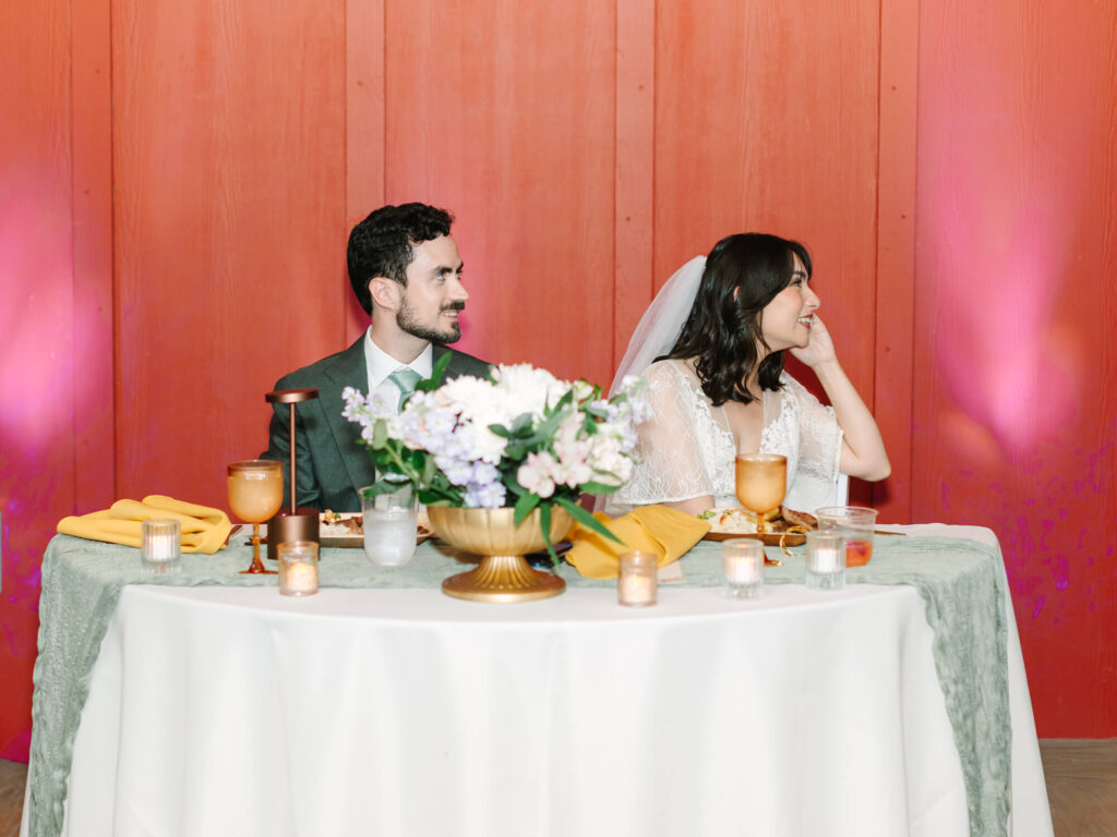 A bride and groom sit at a round table with a white tablecloth, set with candles and flowers.