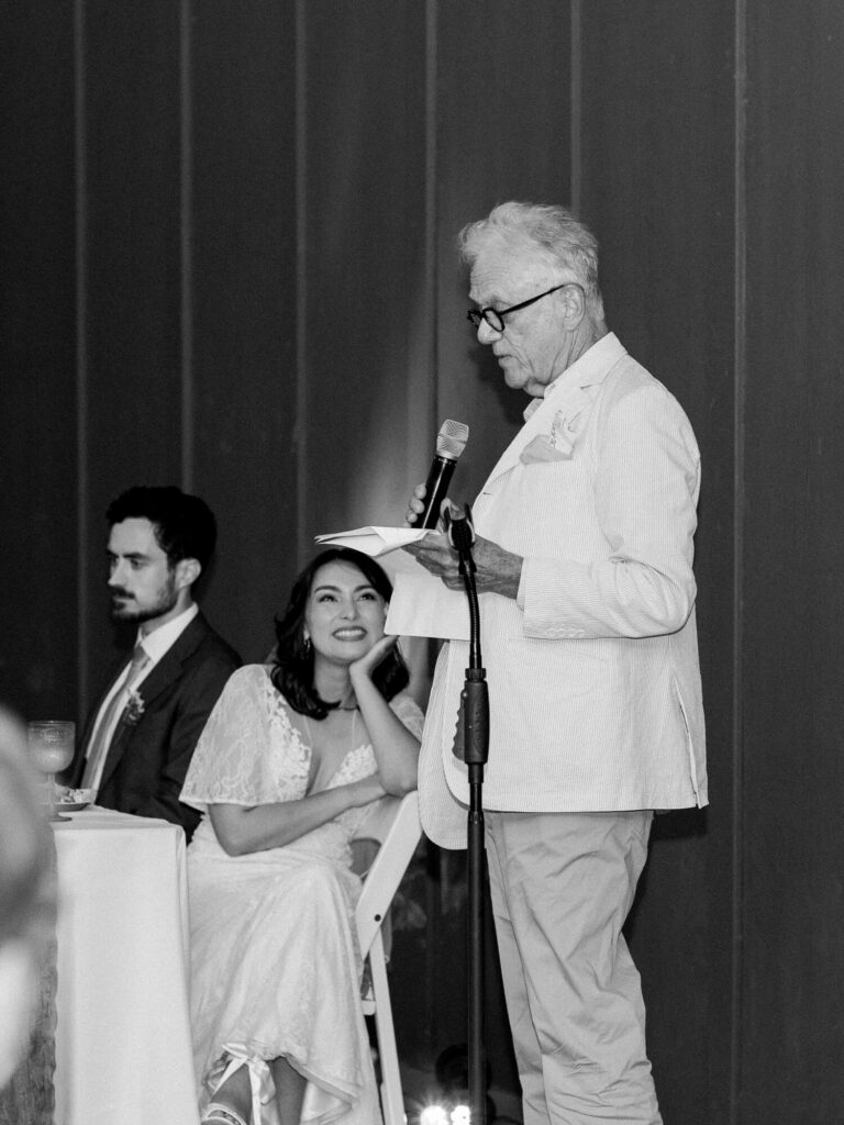A bride smiles warmly at a speech-giver during a wedding reception. A man in a suit sits beside her, and the elder speaker holds a microphone.