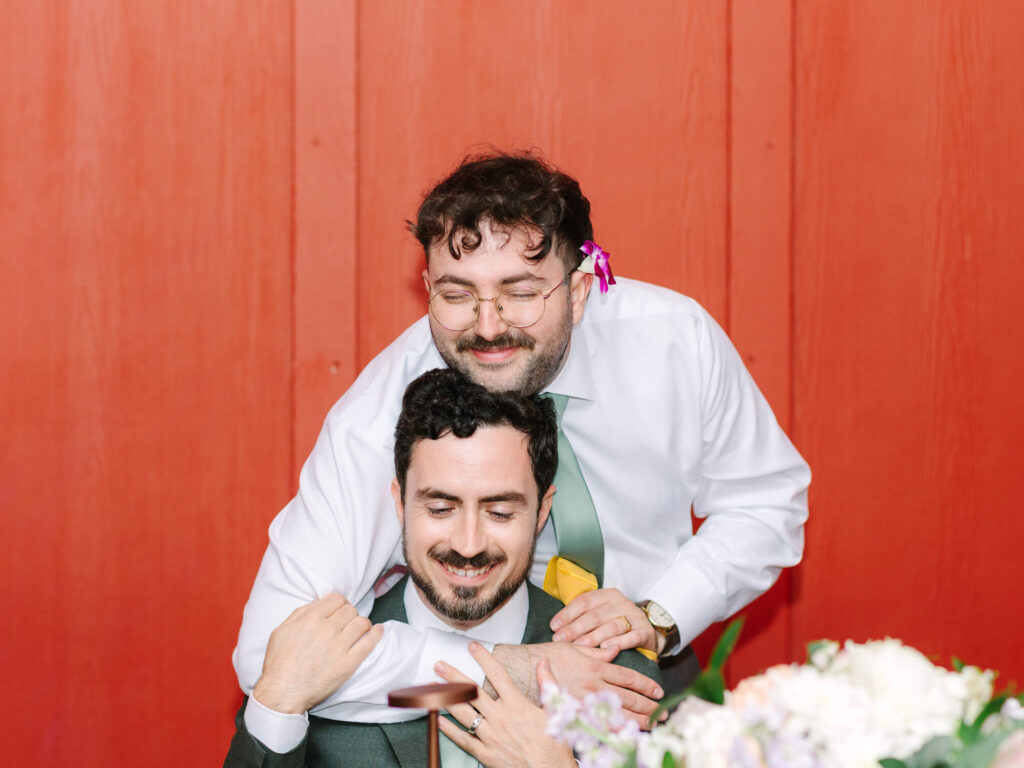 Two men share a joyful embrace in front of a vibrant red background. One wears a flower tucked behind his ear.