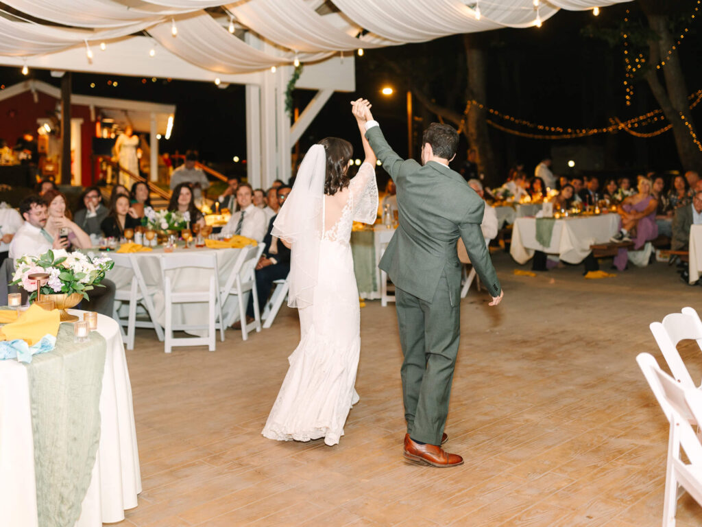 Bride in white gown and groom in green suit celebrate first dance at outdoor evening reception. Guests watch with smiles under string lights and draped fabric.