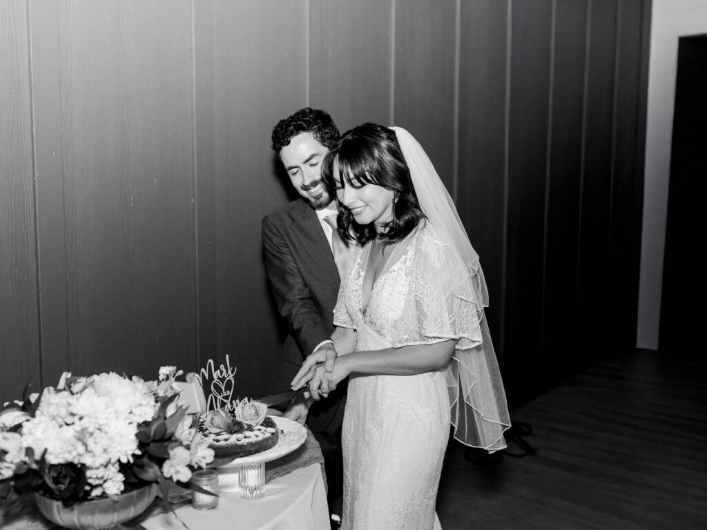A bride and groom joyfully cut their wedding cake together. The bride wears a lace gown and veil, both smiling.