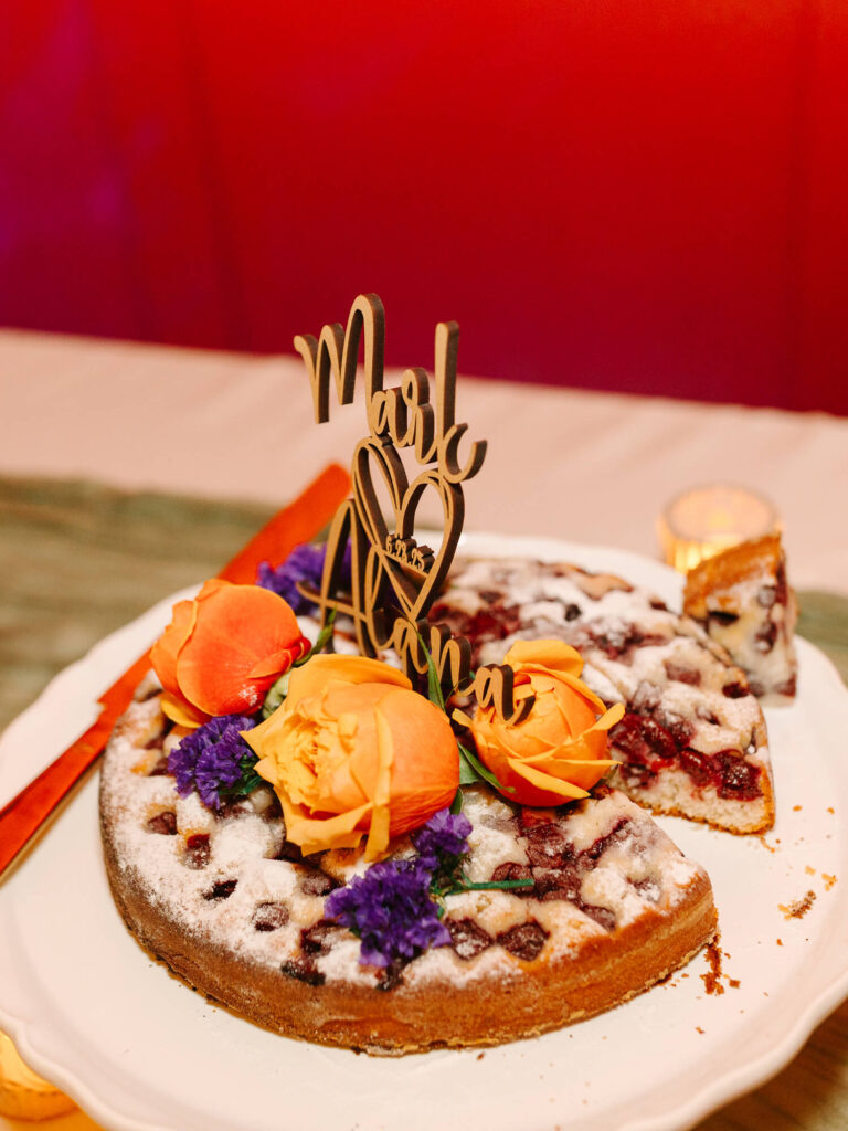 Fruit tart with a heart-accented topper, decorated with vibrant orange roses and purple flowers on a white stand.
