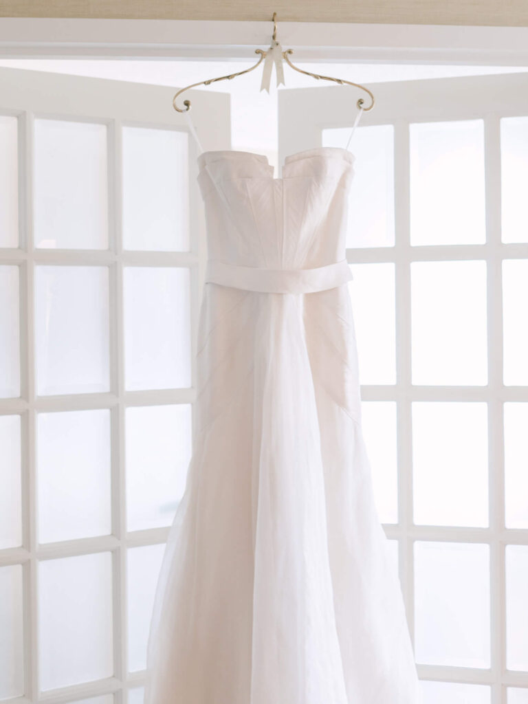 Elegant white strapless wedding dress hanging on a decorative hanger against bright paneled windows.