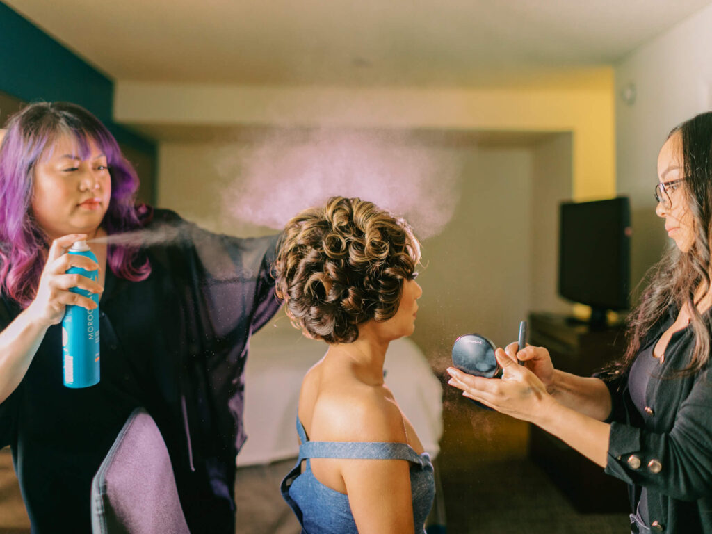 A bride in a blue dress gets her hair styled with hairspray, creating a mist, while another applies makeup.