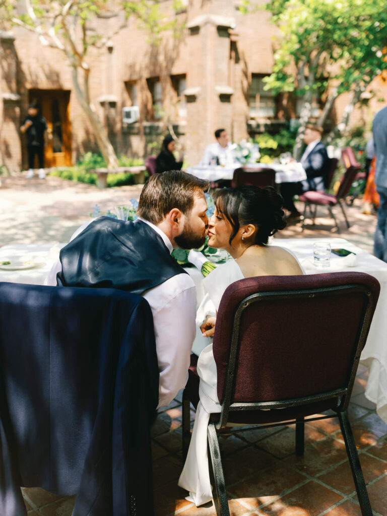 A bride and groom share a tender kiss while seated at an outdoor reception.