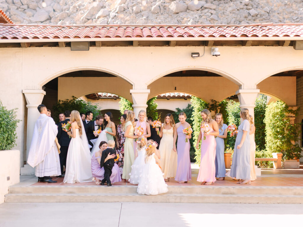 A wedding party gathers under arches in a sunlit courtyard. Bridesmaids in pastel dresses hold bouquets.