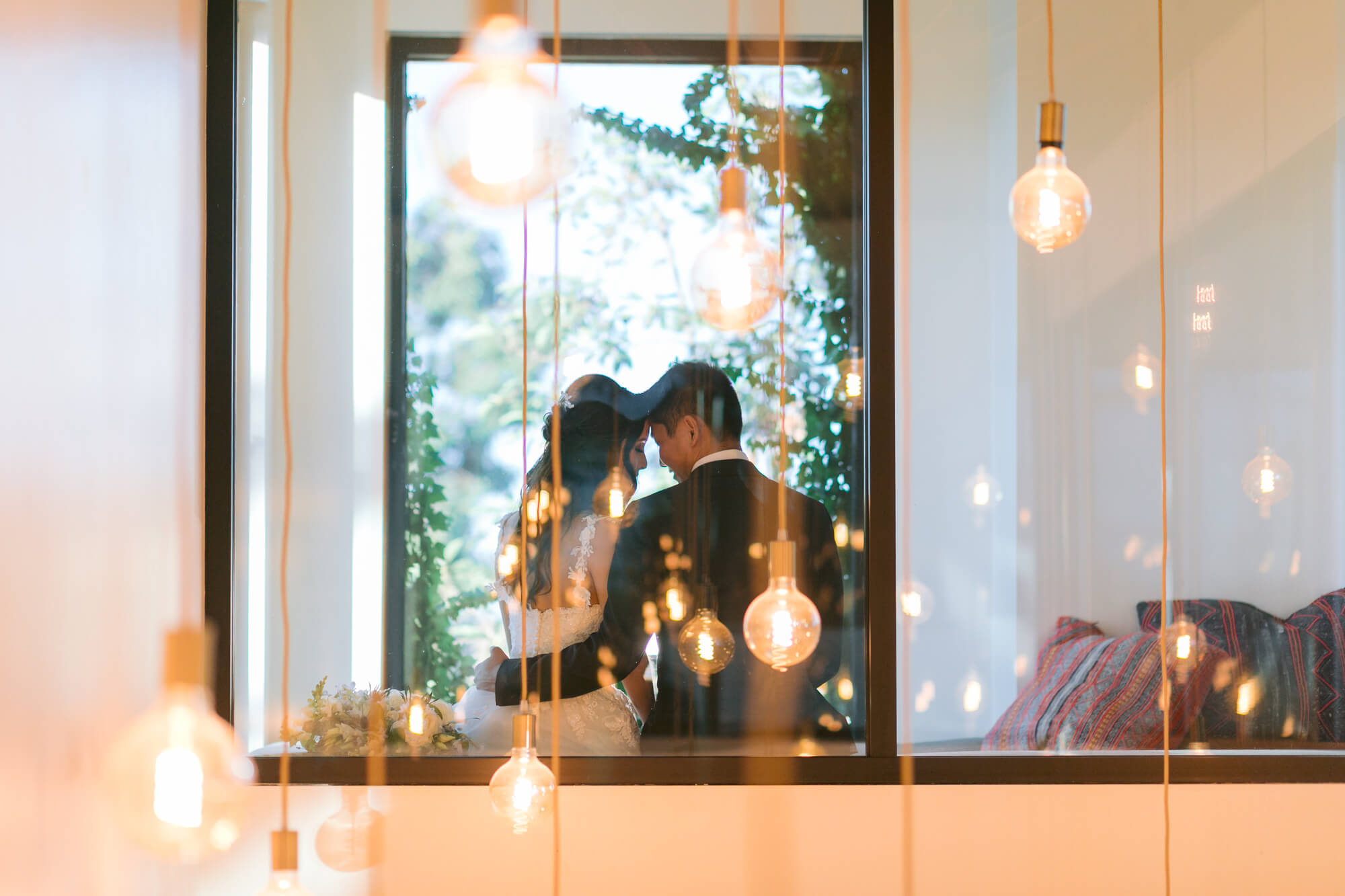 Bride and groom embrace by a window, surrounded by glowing hanging bulbs.
