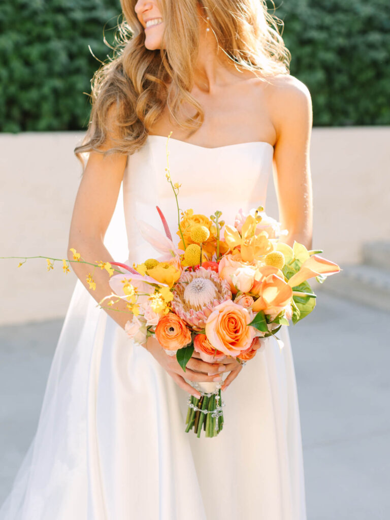 A smiling bride in a strapless white gown holds a vibrant bouquet of yellow and orange flowers. Soft sunlight casts a warm glow, with greenery behind.