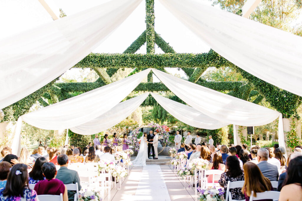 A wedding ceremony under a lush, green arbor adorned with flowing white drapes. Guests seated in rows, overlooking a couple at the altar.