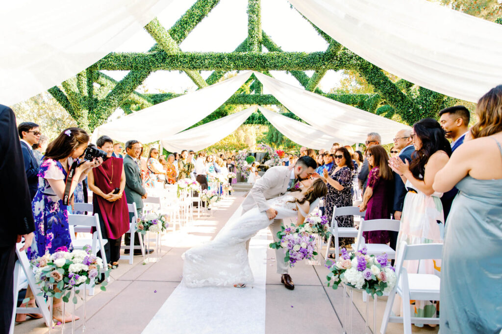 A newlywed couple shares a kiss at the end of an outdoor aisle, surrounded by clapping guests.