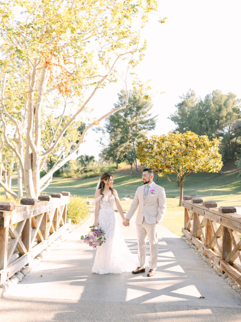 Bride in a lace gown and groom in a beige suit holding hands on a sunny bridge.