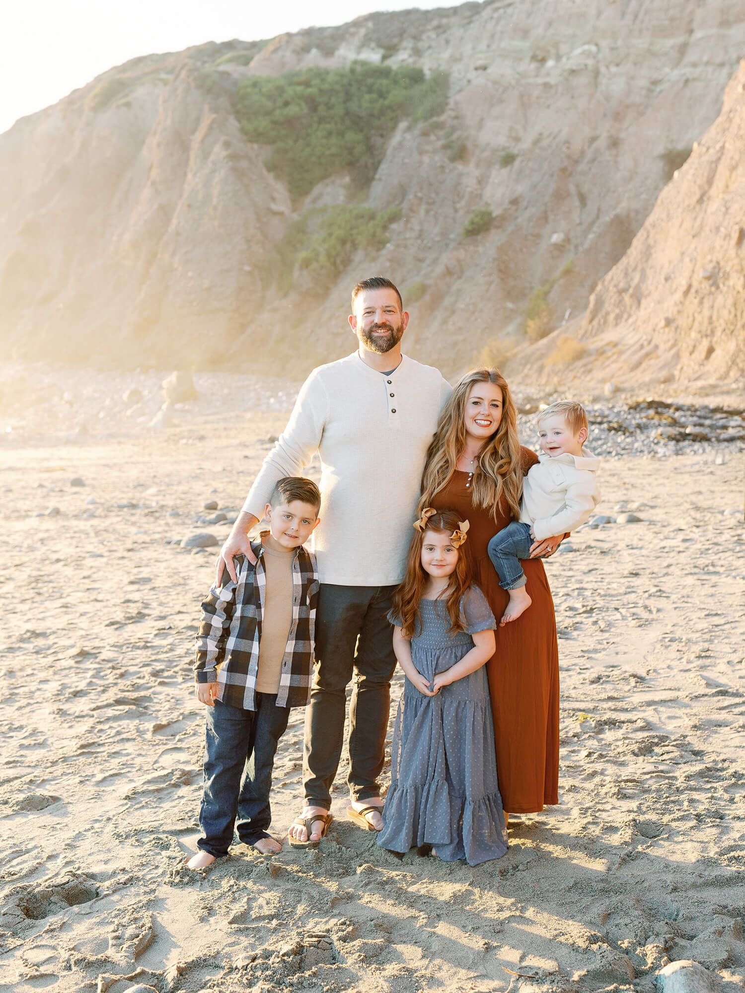 A family of five poses happily on a sunny beach with rocky cliffs in the background.