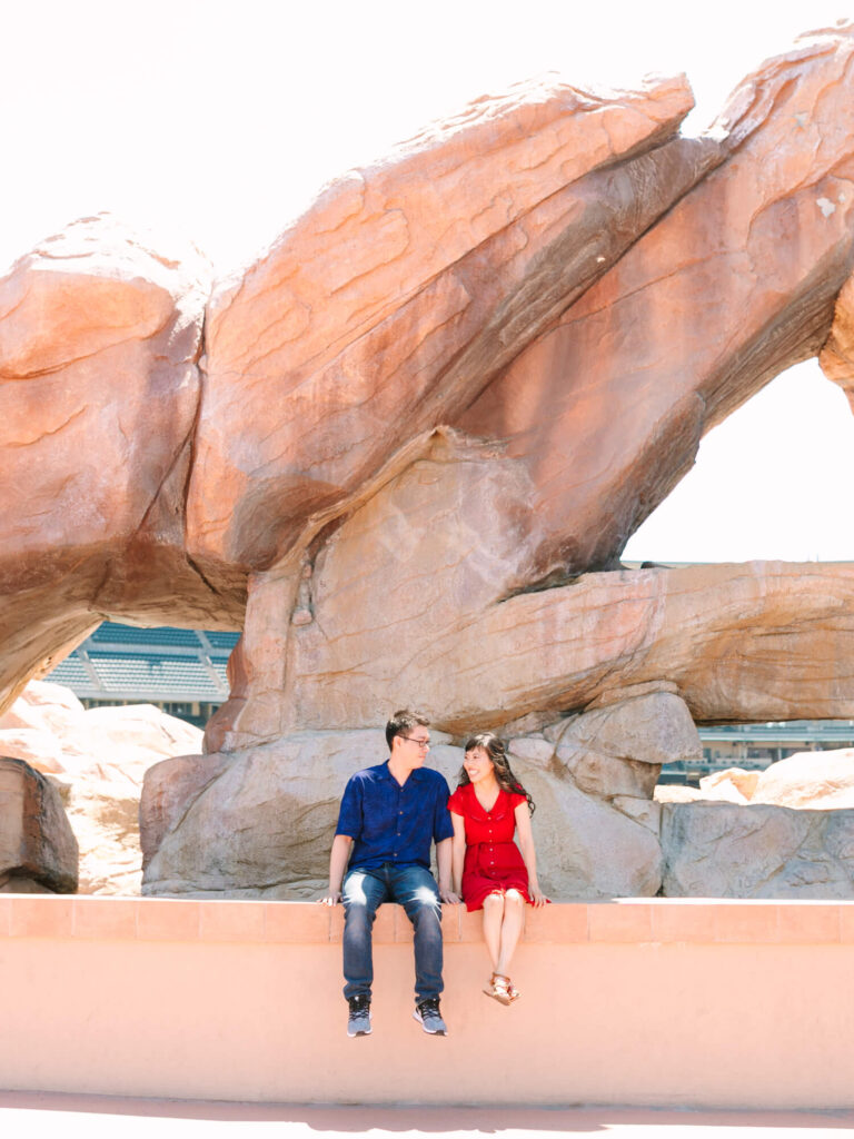 A couple sits on a ledge in front of large, sunlit rock formations. The man wears a blue shirt and jeans; the woman wears a red dress.