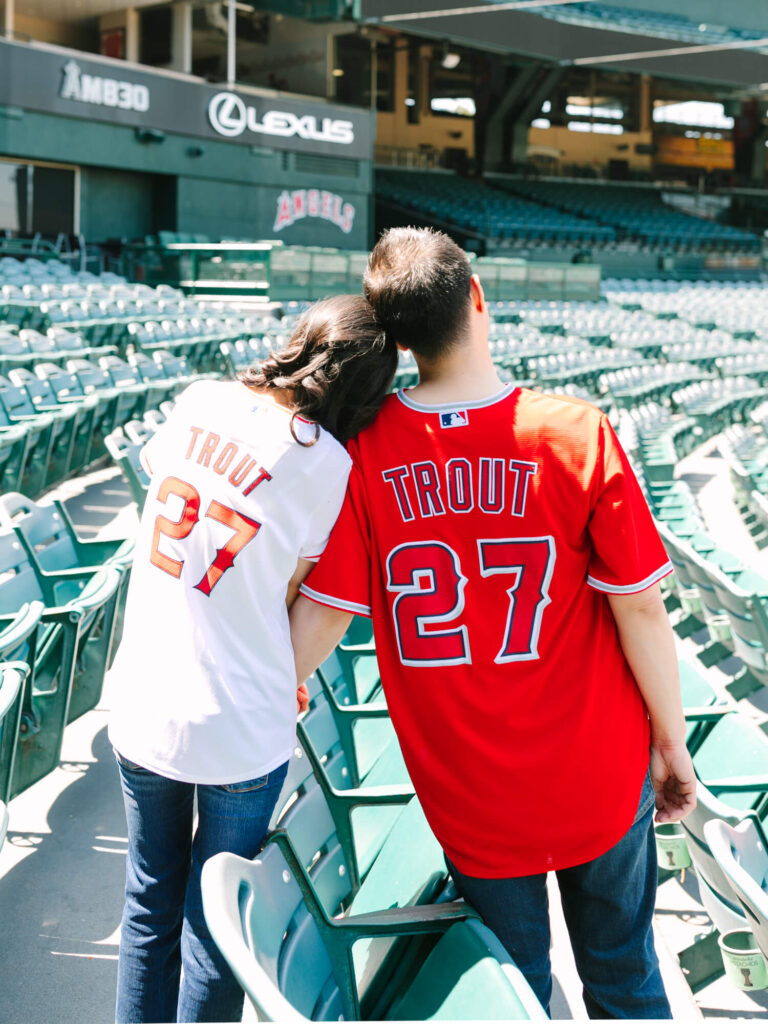 A couple in matching jerseys with "Trout 27" sits in a baseball stadium. The woman leans her head on the man's shoulder.