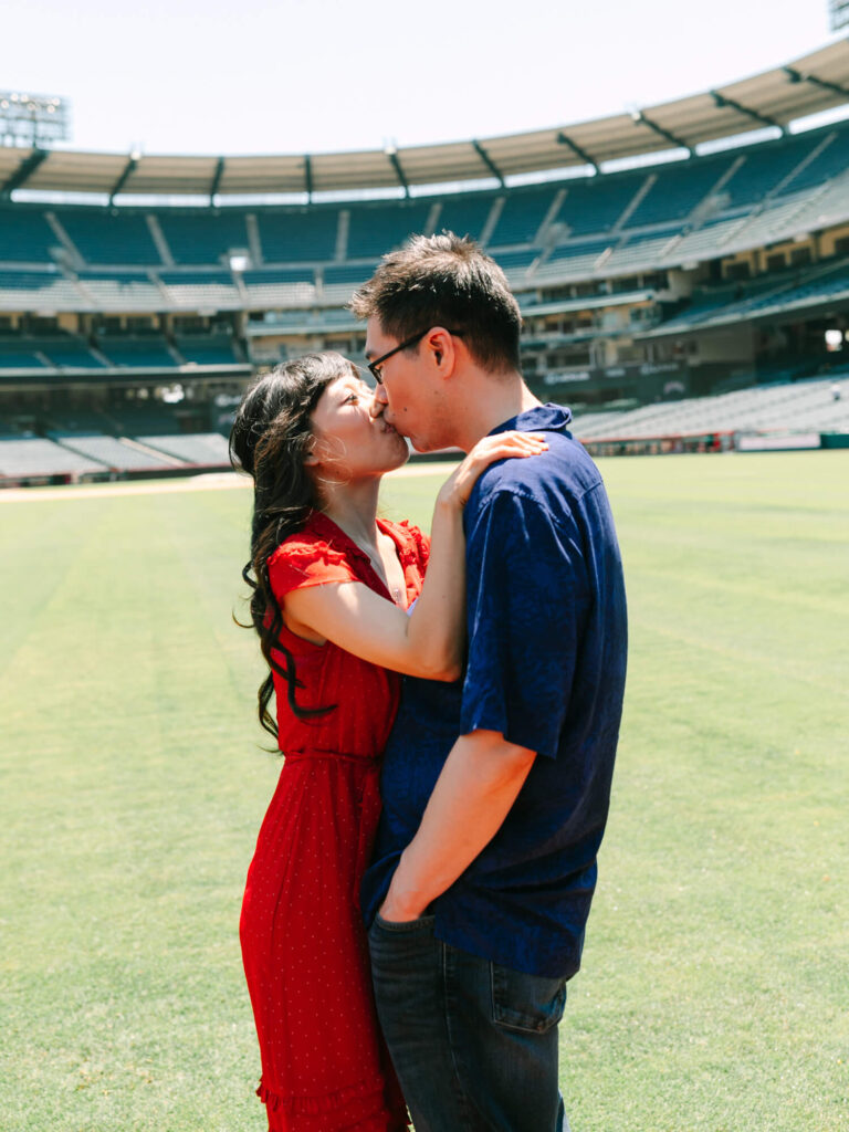 A couple shares a tender kiss on a baseball field. The woman in a red dress embraces the man in a blue shirt.