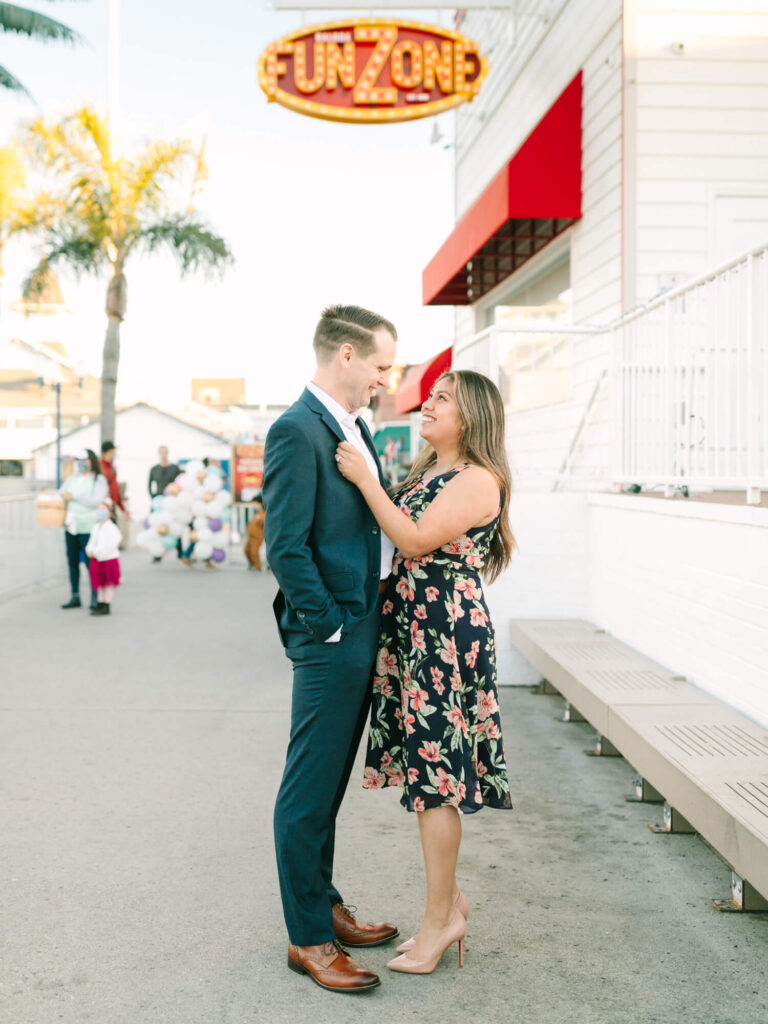 A couple stands outside smiling at each other under a "Fun Zone" sign. The woman wears a floral dress, the man a suit.