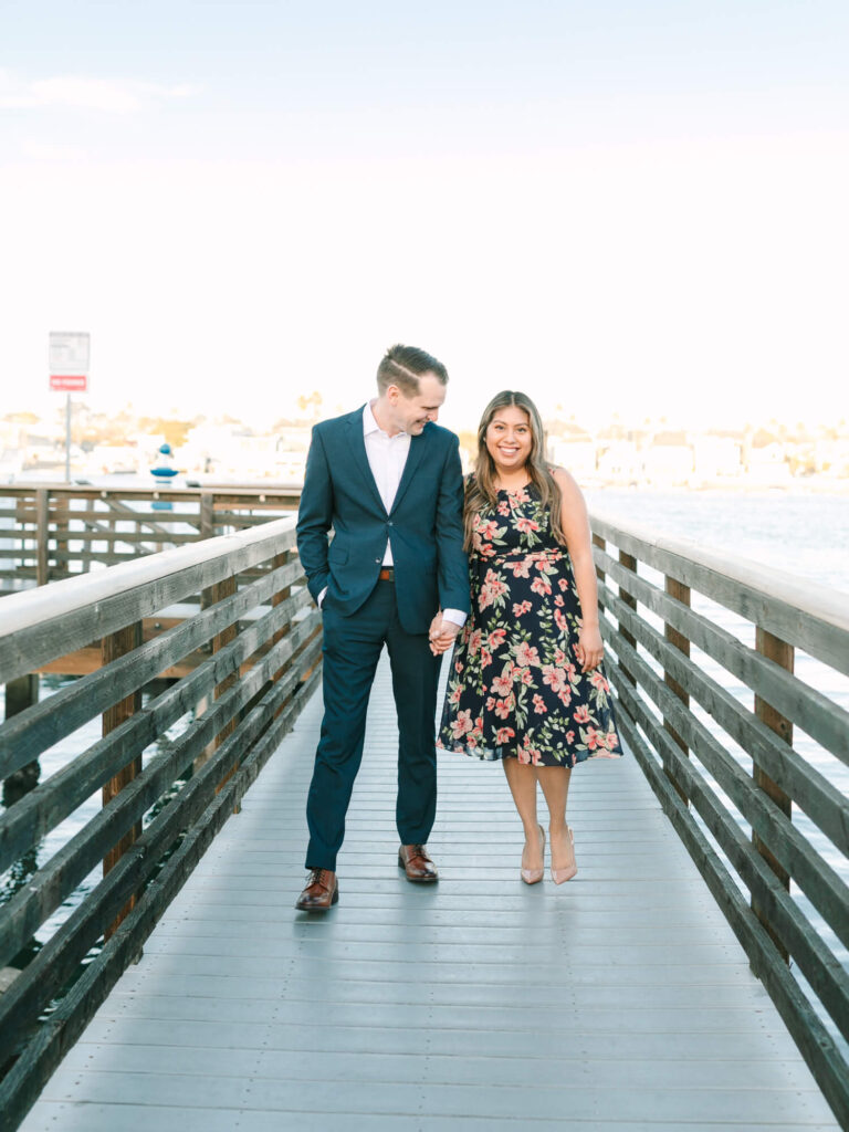 A couple holds hands while walking on a wooden pier. He wears a blue suit; she sports a floral dress.