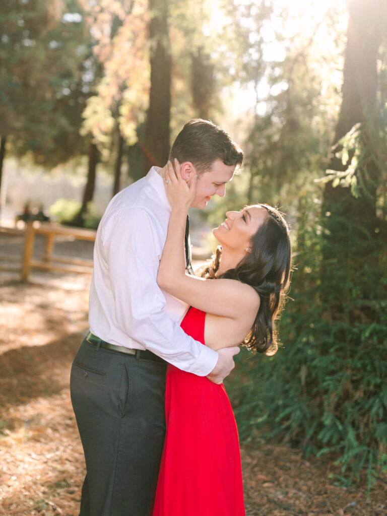 A couple lovingly embraces in a sunlit forest setting. The woman in a red dress gazes up smiling at the man.