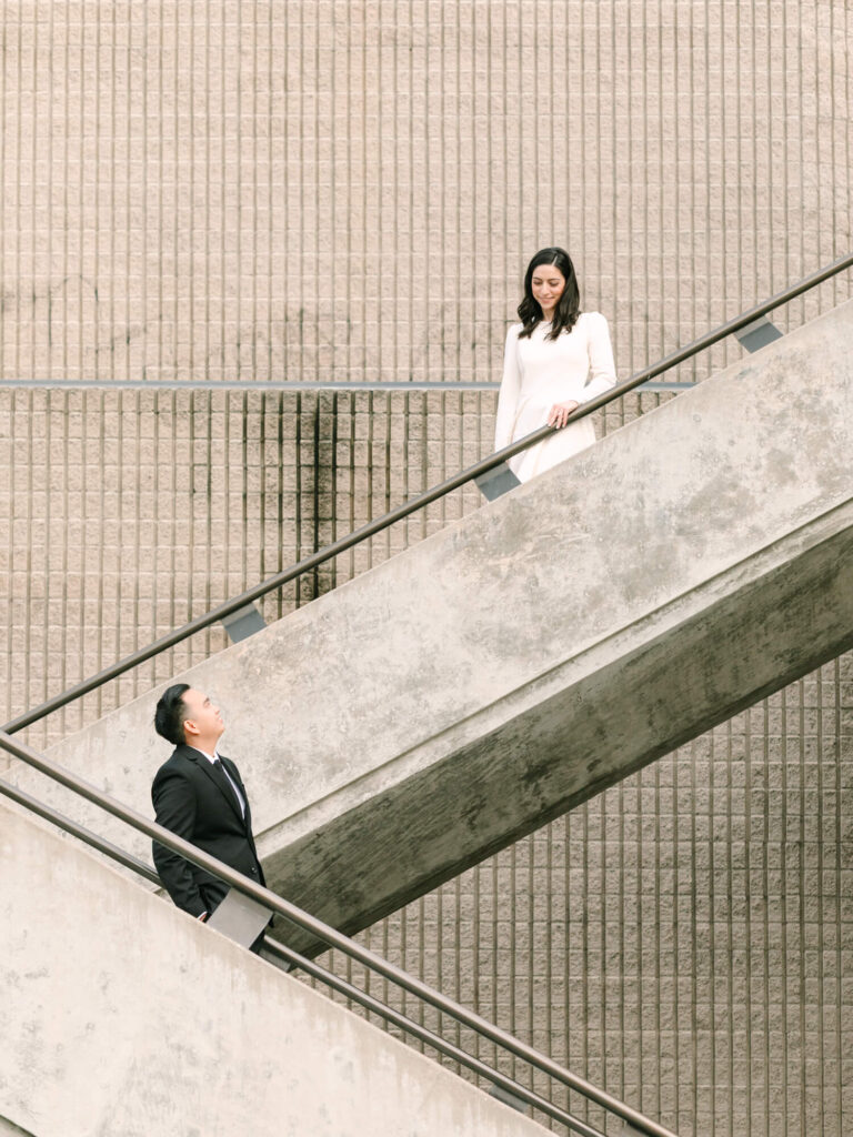 A man in a black suit and a woman in a white dress gaze at each other on a concrete staircase.