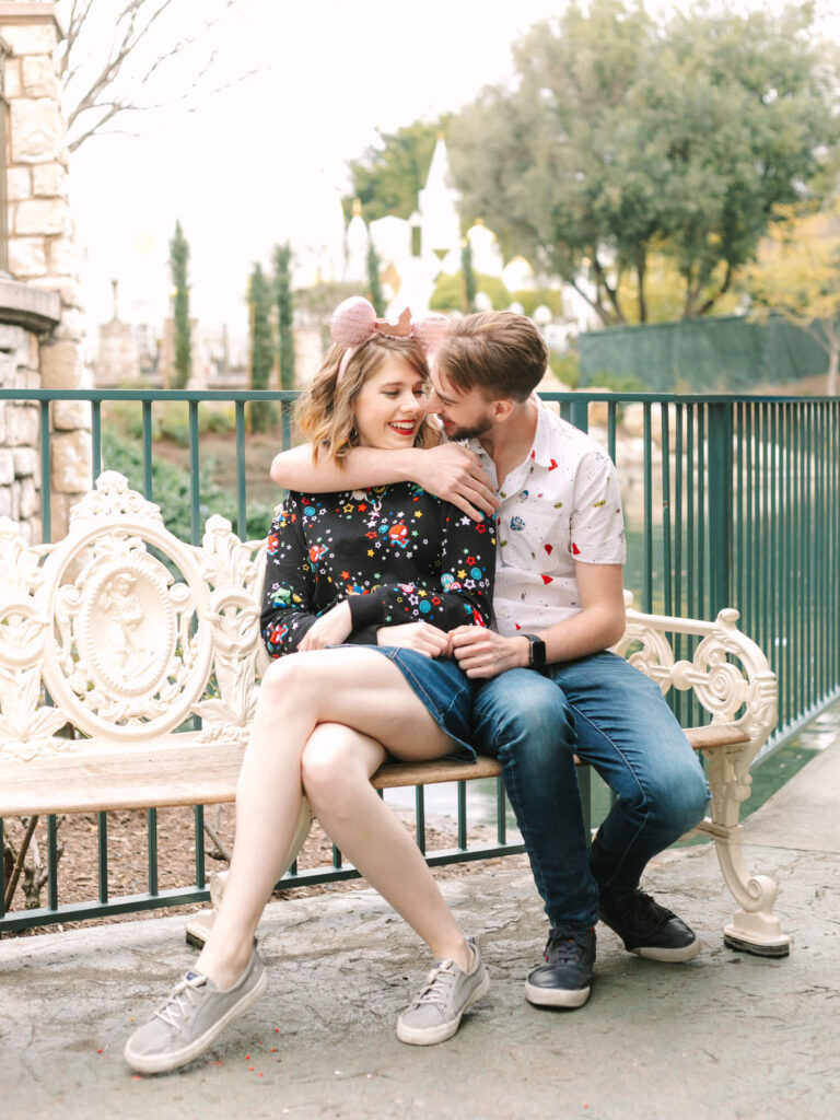 A couple sits joyfully on an ornate park bench, with the woman wearing pink Minnie Mouse ears and a floral sweater.