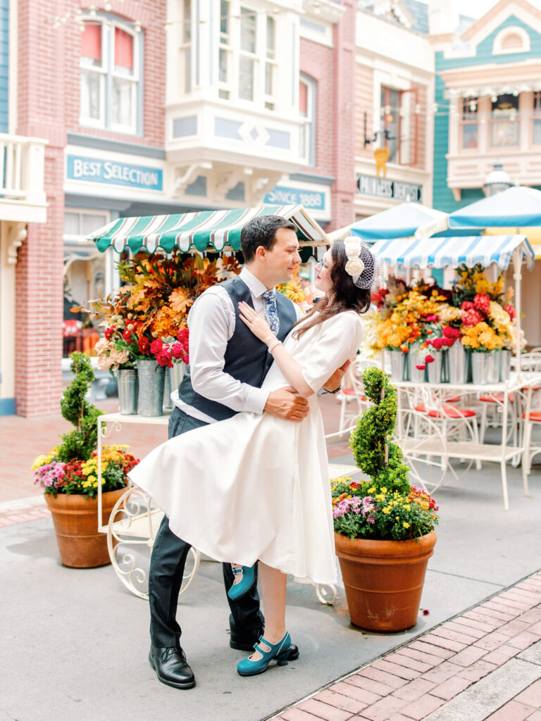 A joyful couple dances on a charming street with floral displays. The woman wears a white dress and fascinator, the man a vest.
