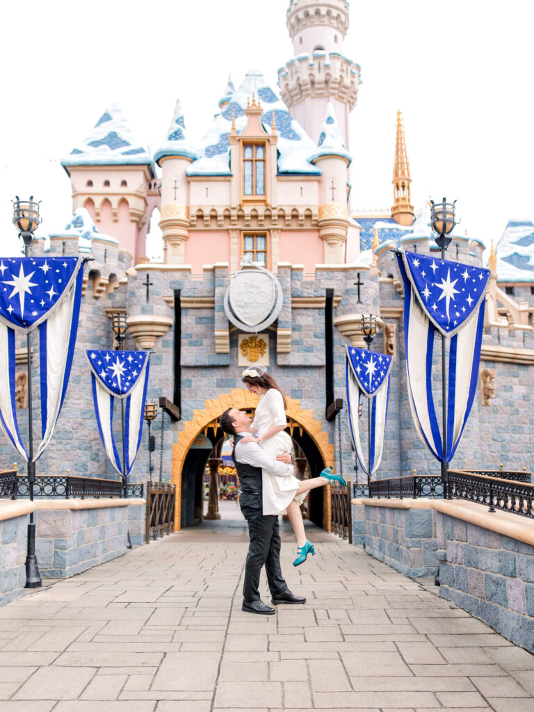 A couple shares a joyful embrace in front of a fairy tale Disneyland castle adorned with blue and white banners.