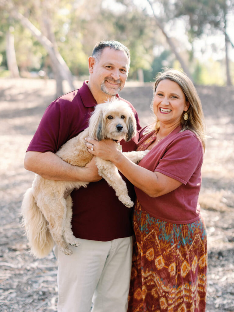 A smiling couple stands outdoors holding a small dog. They wear matching maroon shirts.
