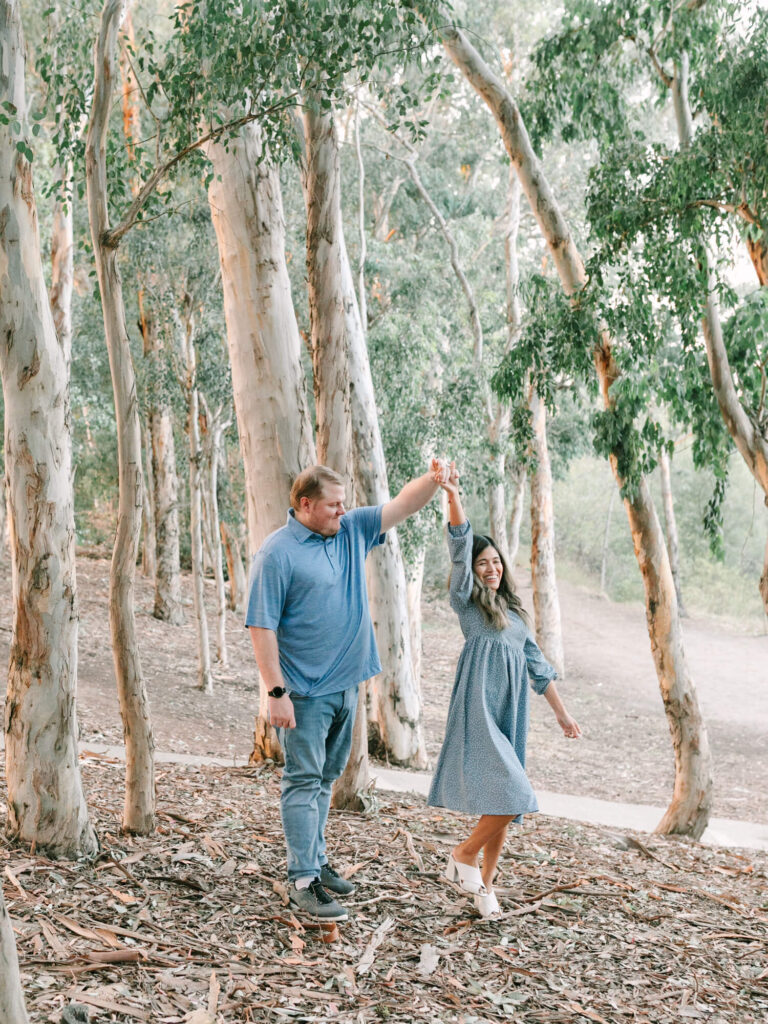 A man and woman happily dance among tall eucalyptus trees. She twirls in a blue dress.